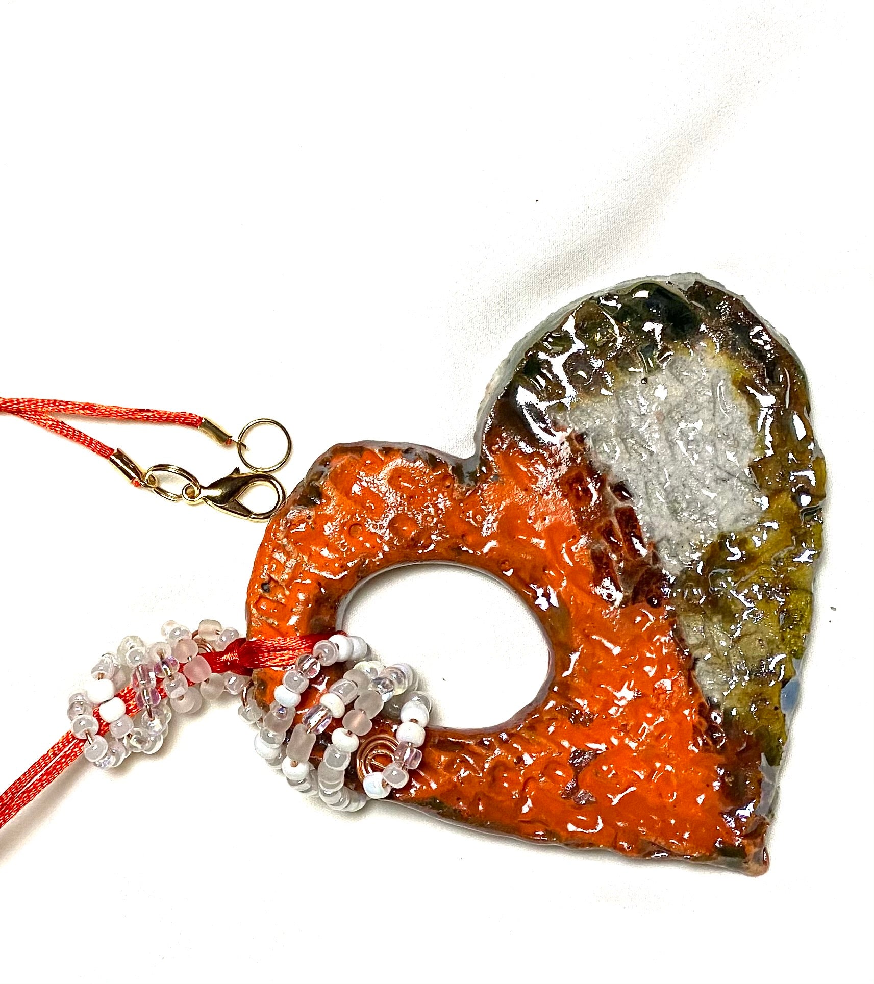 Have A Heart ! Each heart pendant is handmade with love! It is 3"x 3"and weighs approx. 3ozs. This pendant has a red violet and gold metallic raku glazes that renders a unique translucent  patina. The heart has a textured pattern . Both sides are  are different and equally beautiful! It holds a spiral of white and clear mini beads on a spiral copper wire. This pendant has a nice 12" red rattail cord!