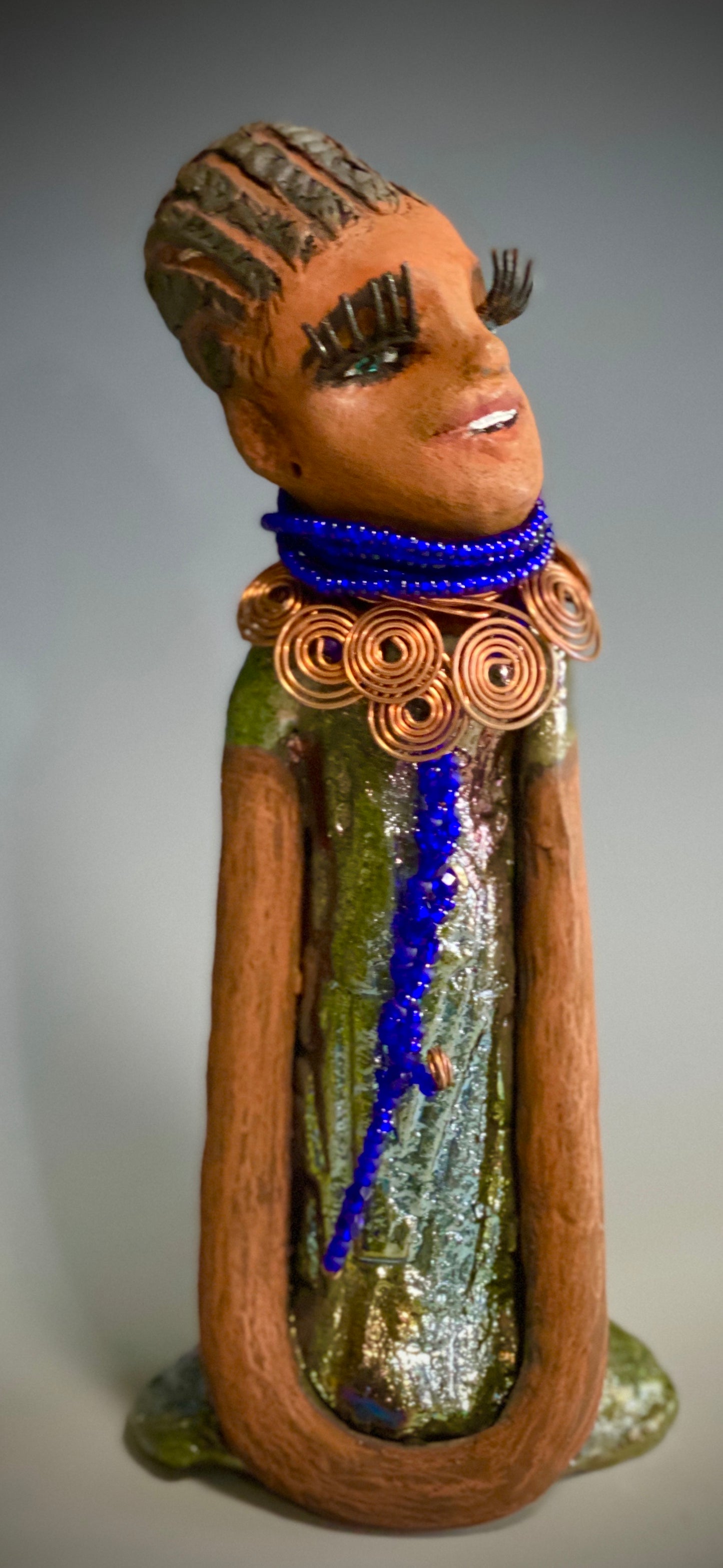 Meet Ophelia! Ophelia stands 7.5" x 4" x 5" and weighs 1.02  lbs. She has a lovely honey brown complexion with reddish brown lips. She has a short braided hairstyle.  Ophelia has a colorful metallic antique copper glazed dress. She wears spiral copper wire necklaces on top of an aqua blue beaded collar. Ophelia long loving arms rest at her side. With accent blue eye shadow and eyes wide opened, Ophelia has hopes of finding  her way into your home!