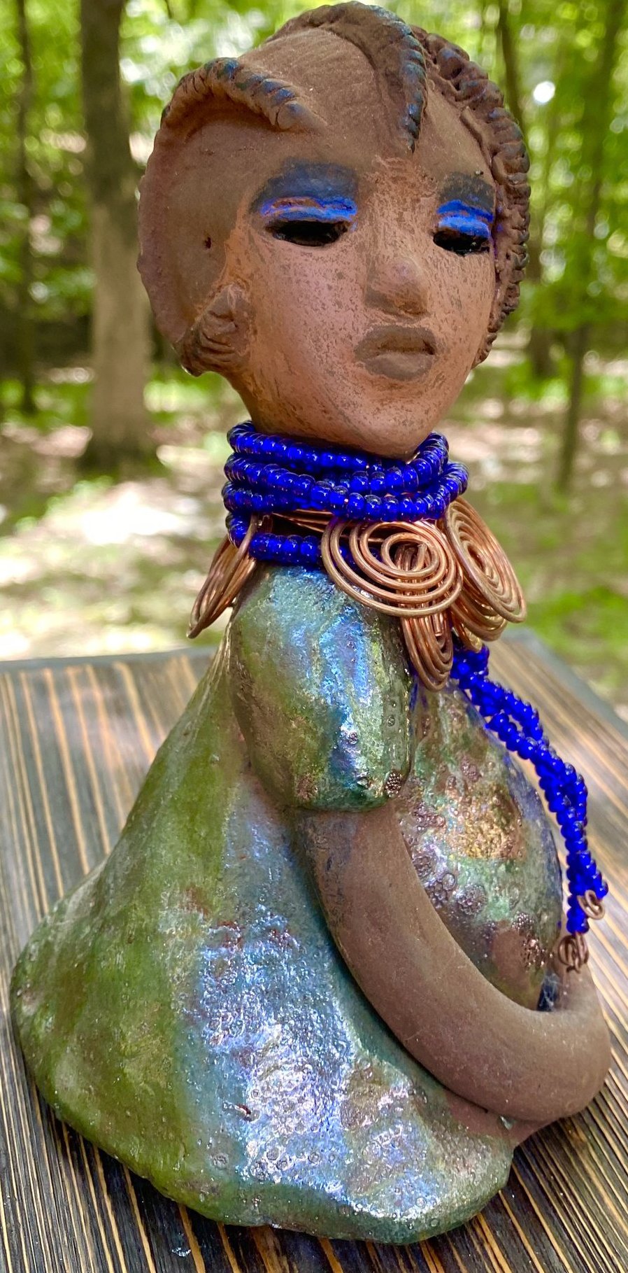 Meet  Ife! Ife stands 7.5" x 4" x 5" and weighs 1.02  lbs. She has a lovely honey brown complexion with reddish brown lips. She has a short braided hairstyle.  Ife has a colorful metallic antique copper glazed dress. She wears spiral copper wire necklaces on top of an aqua blue beaded collar. Ife long loving arms rest at her side. With accent blue eye shadow and eyes wide opened, Ife has hopes of finding a new home.