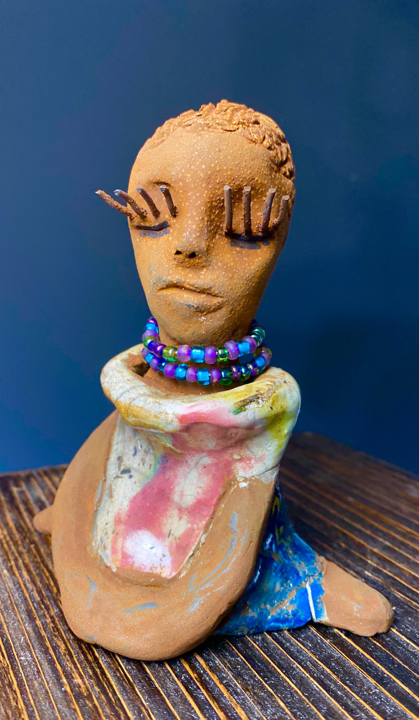 Meet Little Riley! Riley stands 5" x 5”x 3" and weighs  11 ozs.  Riley has a lovely multicolored glossy dress with a matching beaded necklace.  She has long lashes!  Riley appears to sit in a yoga pose. Her long arms rest at her side. Riley is a great starter piece from the Herdew Collection!