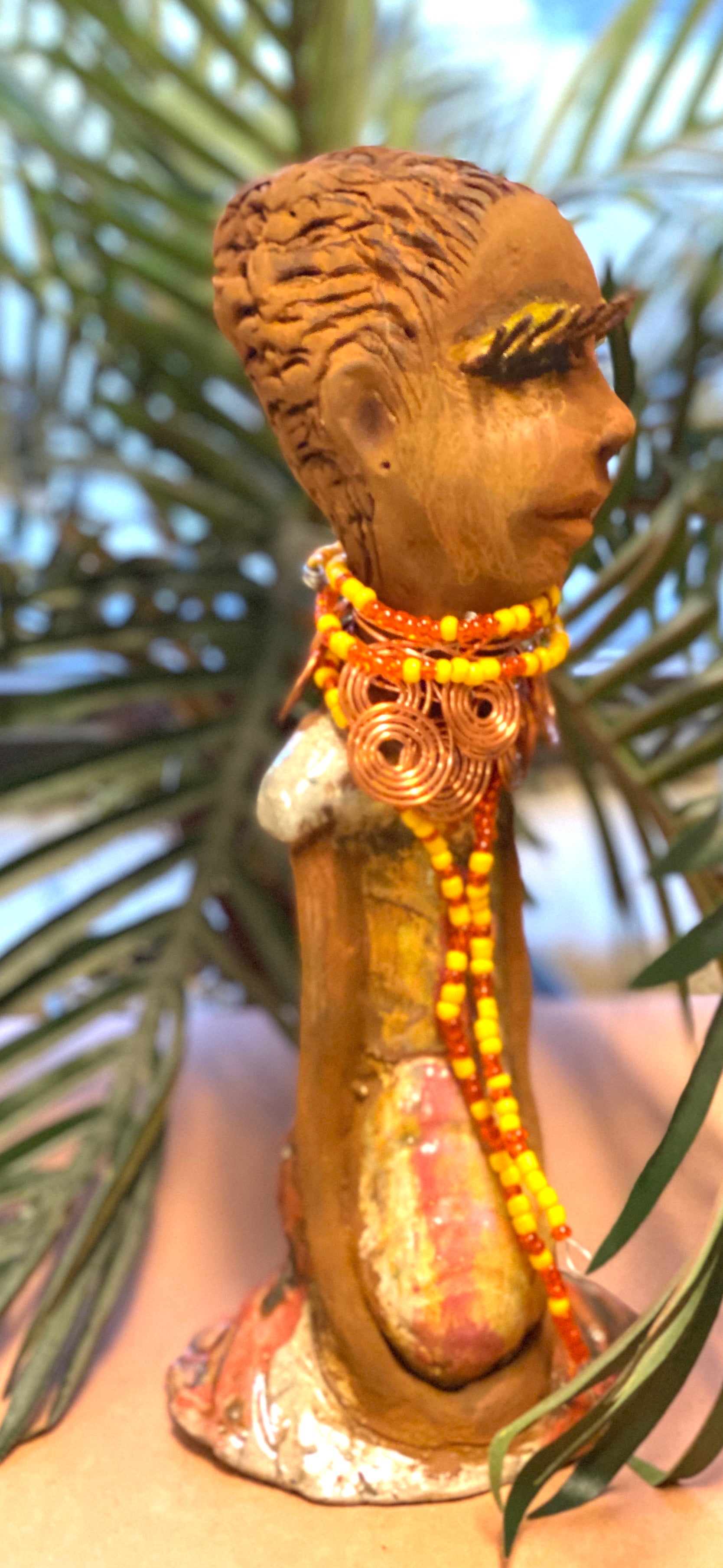 Reandra has a colorful reddish metallic green antique copper glazed dress. She wears a spiral copper wire necklace on top of a yellow red beaded collar. With eyes slightly opened, Reandra has hopes of finding a new home.