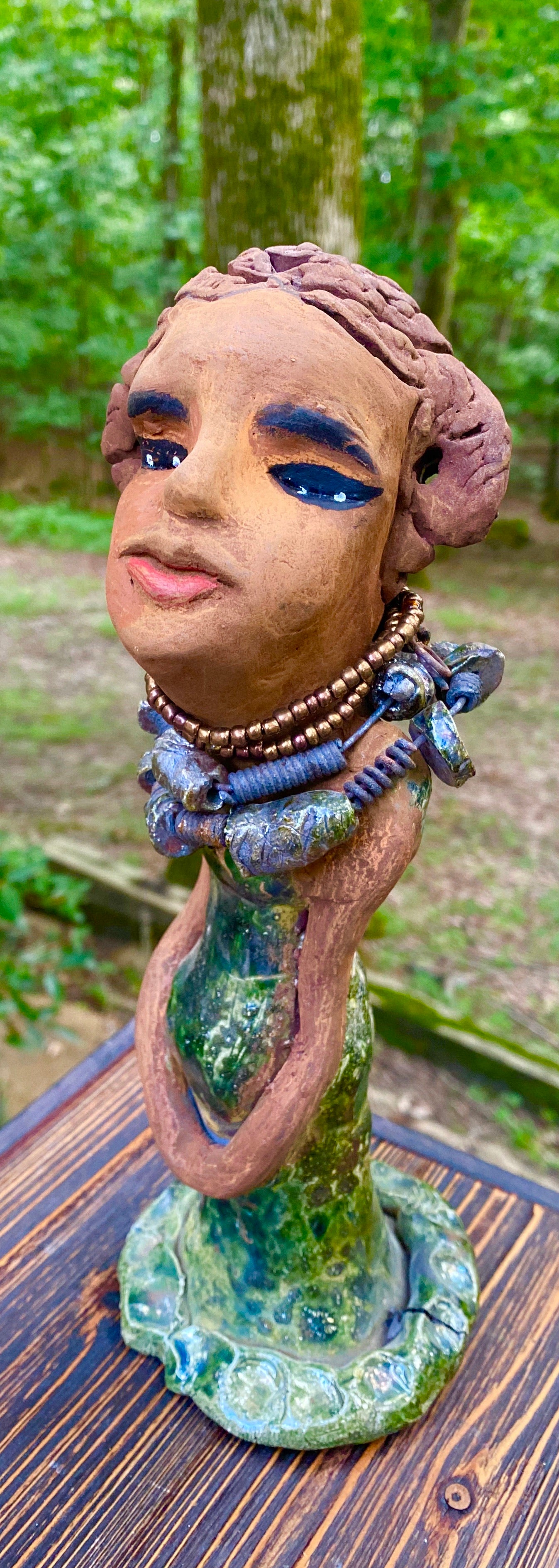 Greta! Greta stands 11" x 4" x 4" and weighs 2.5 lbs. She has a lovely honey brown complexion with  reddish brown lips. She has a braided hairstyle.  Gretaya has a colorful metallic green antique copper glazed dress. She wears two raku beaded necklaces. With long eyes slightly opened, Greta has hopes of finding a new home.